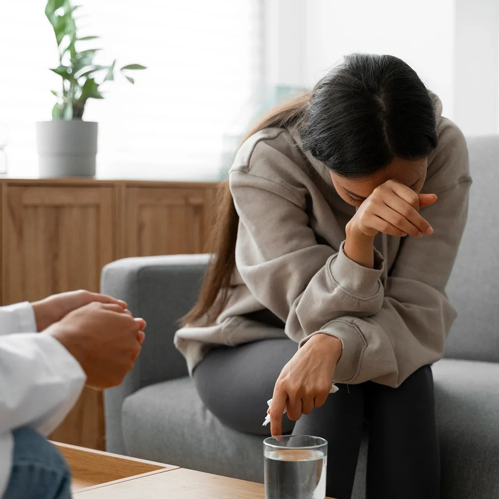 A woman sits in a chair, covering her face with her hand, while a person with a white coat is seated opposite her.