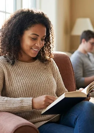 A young woman smiles while reading a book on a couch, with a person in the background.